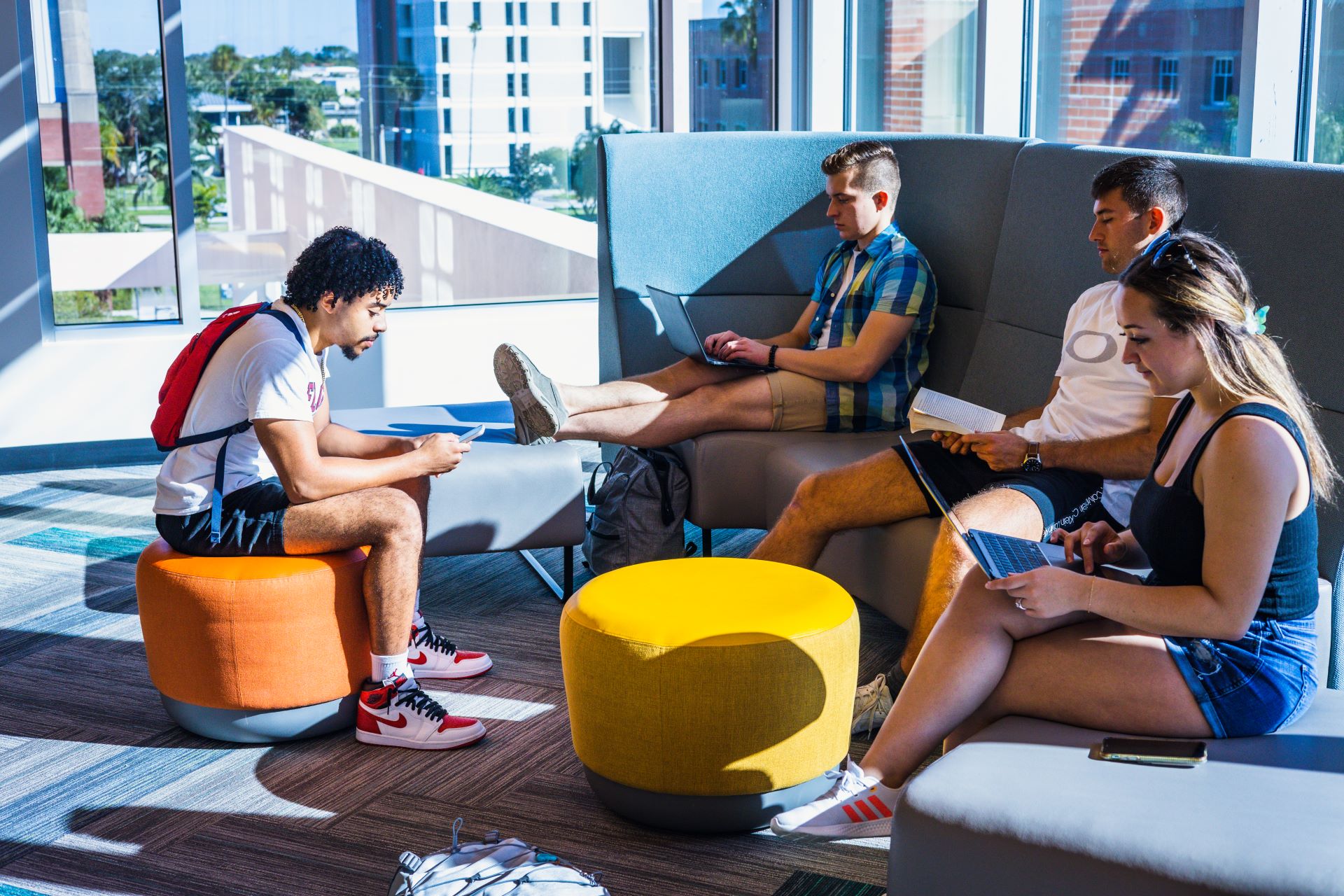 Four students in a lobby, one on a stool and three on a couch, with laptops and books